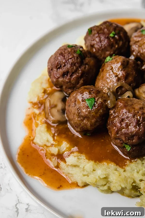 A white plate with mashed cauliflower topped with salisbury steak meatballs, garnished with fresh parsley.