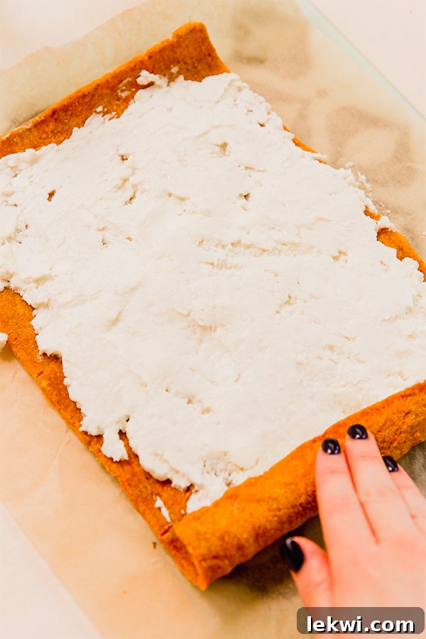 A paleo pumpkin cake unrolled on parchment paper, beautifully frosted with a white, creamy dairy-free icing.