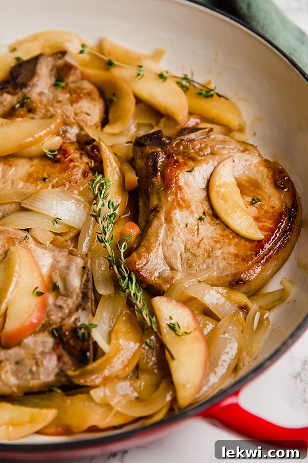 Golden-brown pork chops nestled among caramelized apple slices and onions in a large cast iron skillet, simmering in a rich sauce.