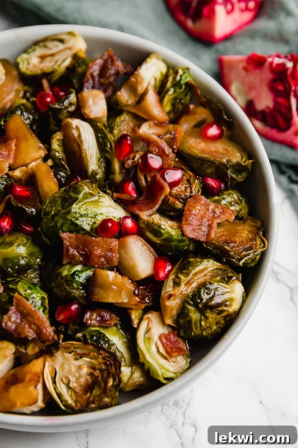 A white bowl filled with perfectly roasted brussels sprouts, tender apple pieces, and crispy bacon bits, garnished with fresh pomegranate seeds.
