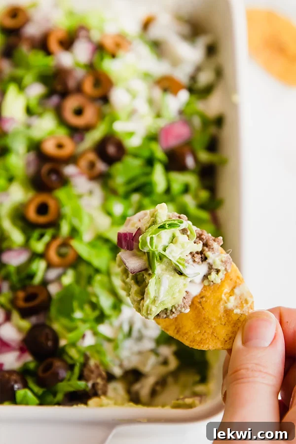 A crisp plantain chip expertly scoops a generous portion of layered paleo taco dip, showcasing the vibrant colors and creamy textures of guacamole, ground beef, and other fresh toppings.