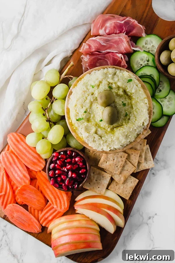 A beautifully arranged wooden charcuterie board showcasing a vibrant assortment of fresh apples, crisp carrots, glistening pomegranate seeds, juicy grapes, gluten-free crackers, creamy hummus, refreshing cucumber slices, and savory cured meats.