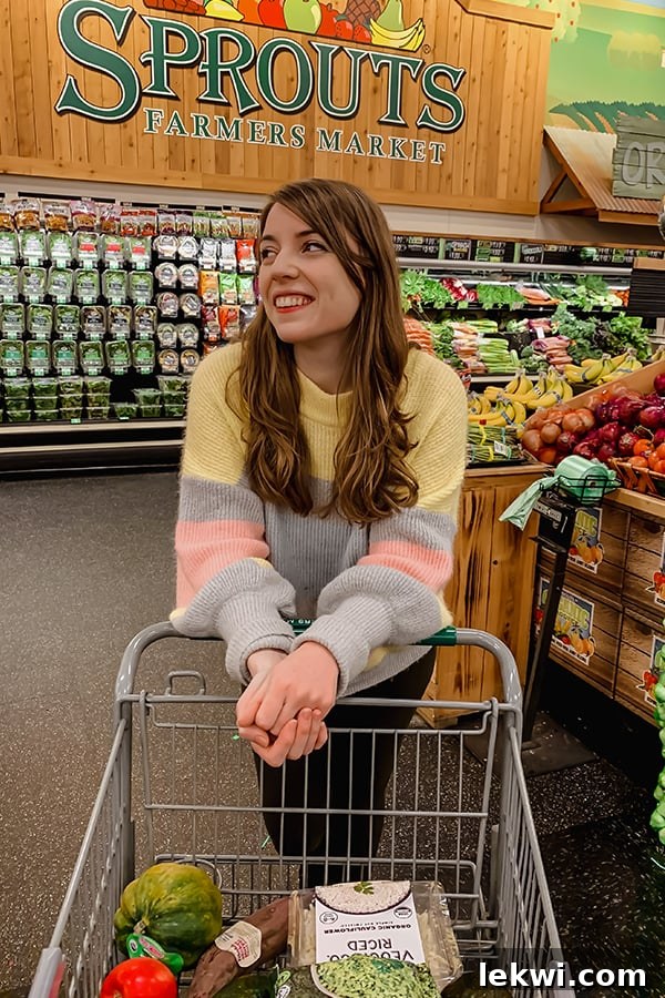 A cheerful woman standing in the vibrant produce section of Sprouts, leaning on a shopping cart, ready to explore healthy options.