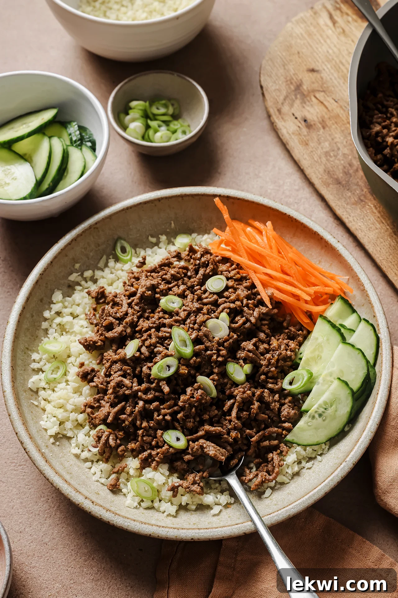 A vibrant Korean beef bowl featuring seasoned ground beef, fresh carrots, crisp cucumber, and fluffy cauliflower rice. Additional toppings are neatly arranged in smaller bowls alongside the main dish, highlighting a colorful and inviting meal presentation.