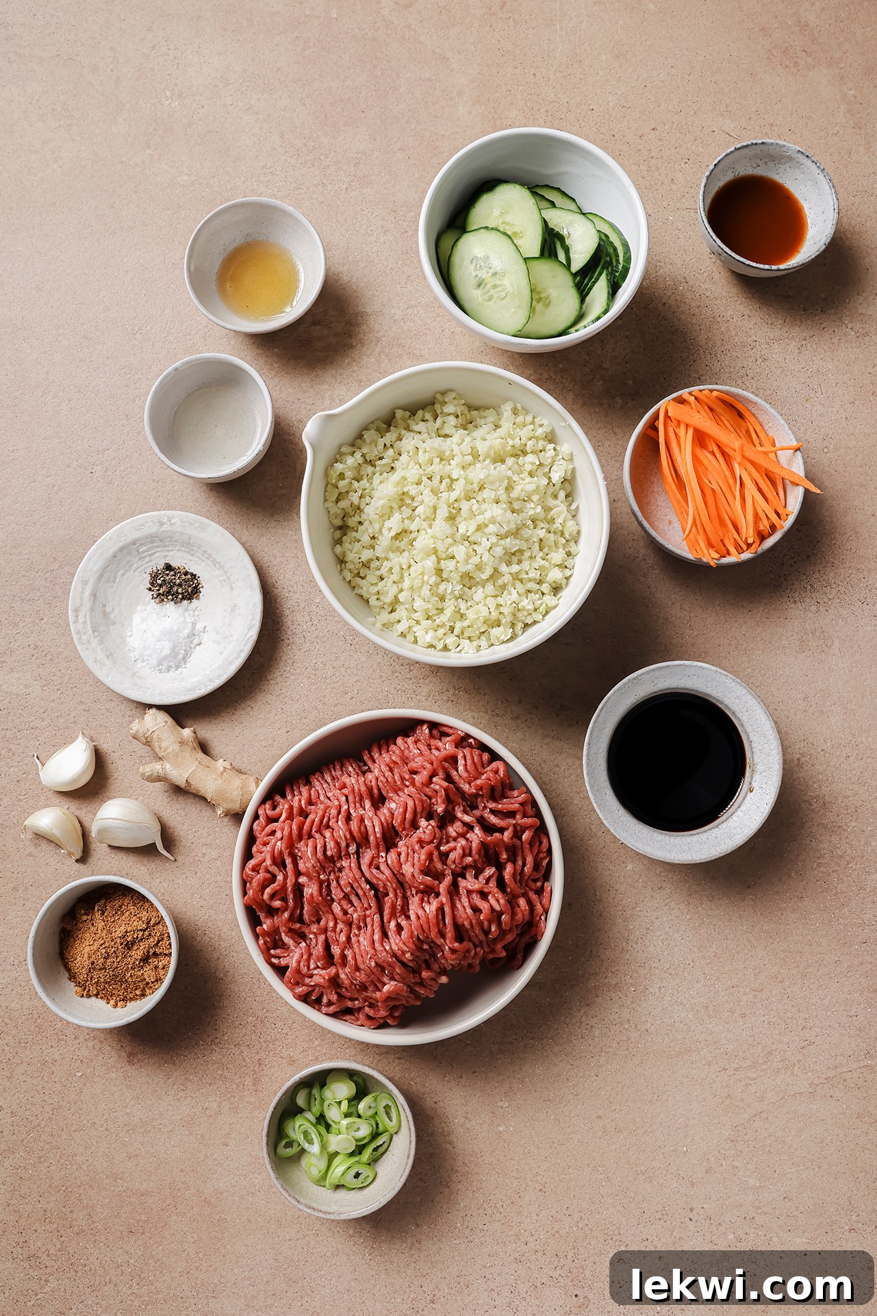 All the fresh ingredients required to prepare Korean ground beef bowls are neatly arranged in small white bowls and on a cutting board, prior to cooking. The array includes ground beef, ginger, garlic, coconut aminos, sriracha, and various vegetables like carrots and cucumber, ready for preparation.