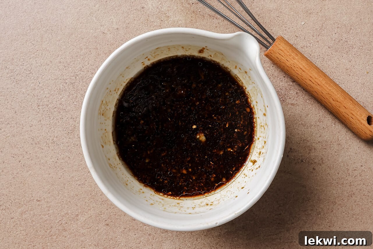 A small white bowl containing a vibrant, reddish-brown sauce is positioned on a light-colored surface next to a metal whisk, suggesting the initial preparation of a dressing or marinade.