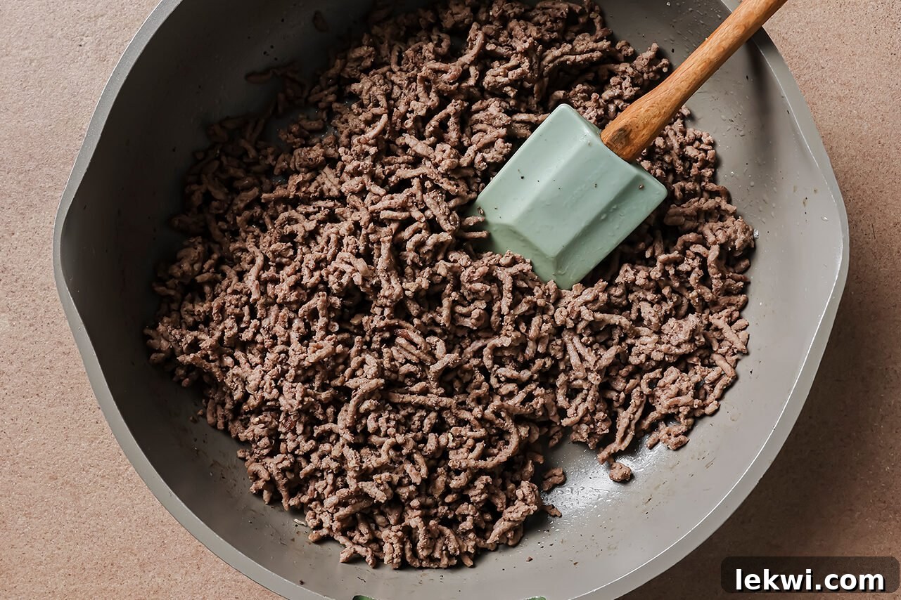 A non-stick sauté pan on a stove, filled with browning ground beef that is being broken apart with a wooden spatula. Minced garlic and grated ginger are visible, mixed in with the beef as it cooks, indicating the aromatic start of the dish.