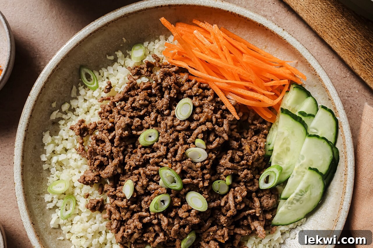 A beautifully assembled Korean ground beef bowl, showcasing the rich, saucy beef as the centerpiece. It's surrounded by vibrant sides of fluffy cauliflower rice, crisp cucumber slices, and bright julienned carrots, all garnished with fresh green onions for a colorful and inviting meal.