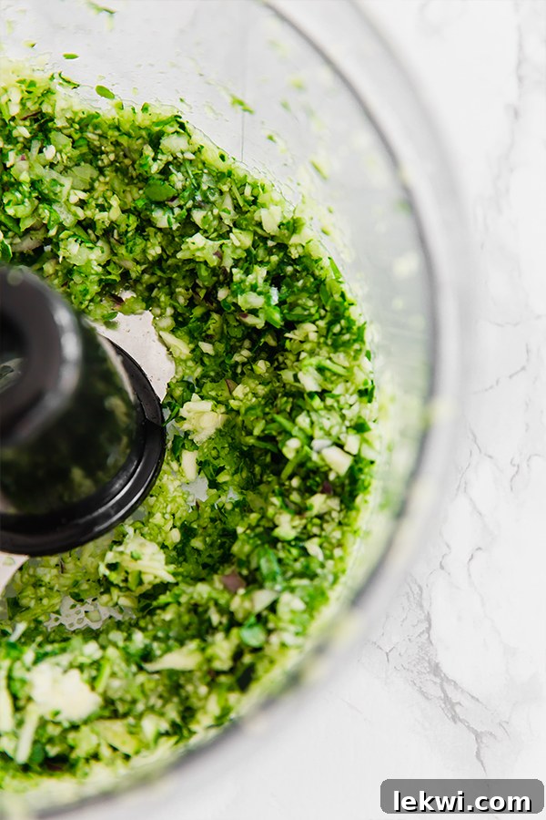 A food processor actively blending fresh herbs and vegetables like spinach, zucchini, red onion, garlic, and parsley, illustrating the first step of finely shredding ingredients for the loaded veggie & chicken meatballs.