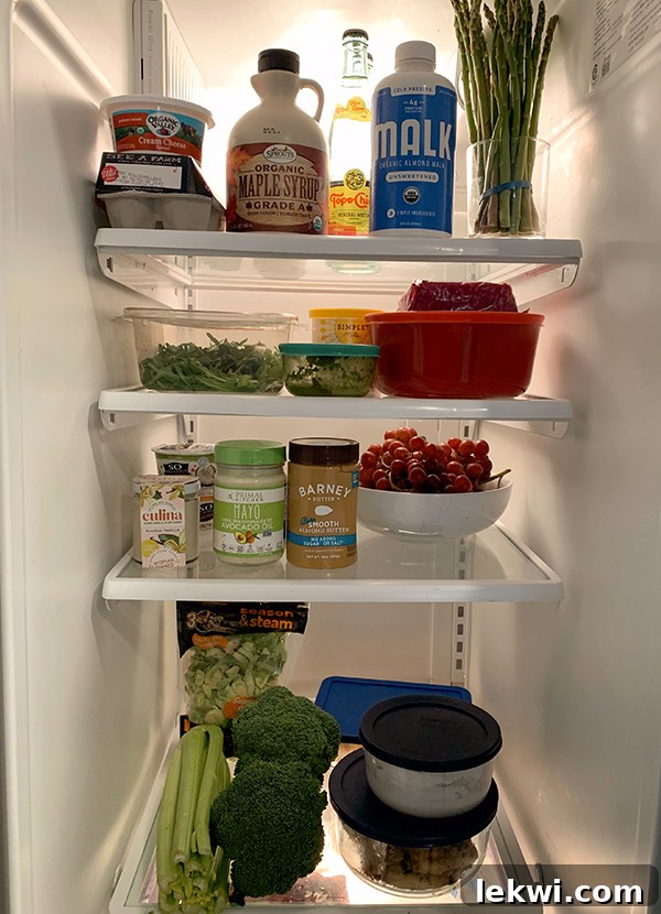 A look into the fridge with vegetables, almond butter, mayo, grapes, tupperware containers, almond milk, and maple syrup. 