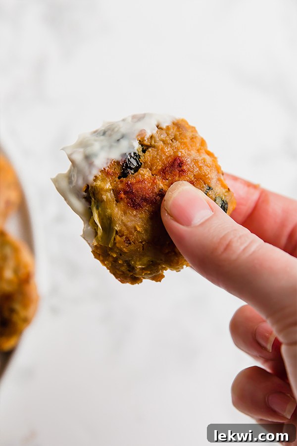 A hand holding a spinach and artichoke sweet potato chicken popper, dipped into a creamy white sauce, highlighting its golden exterior.