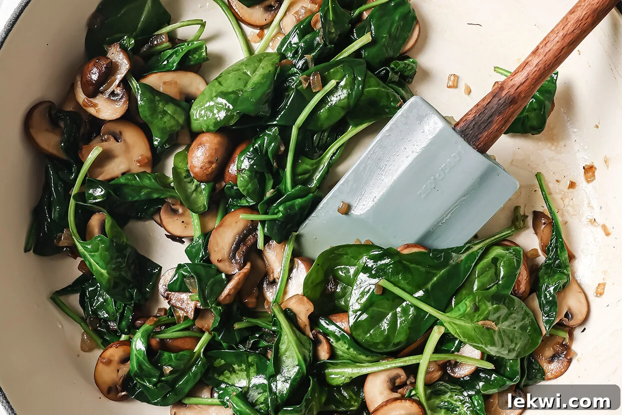 Mushrooms and fresh spinach being cooked in a pan with a spatula, wilting into the sauce.