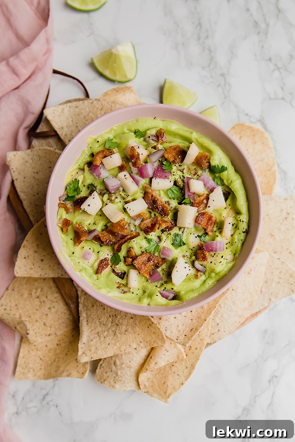 A beautifully presented bowl of Apple Bacon Guacamole, surrounded by various dipping options like plantain chips and vegetable sticks, ready for a party.