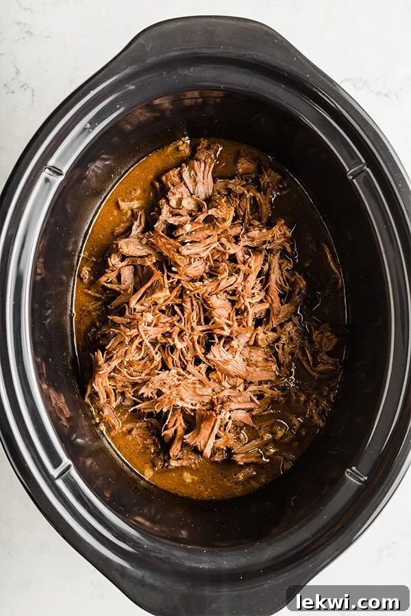 Shredded beef simmering in a slow cooker, surrounded by rich broth and bay leaves.