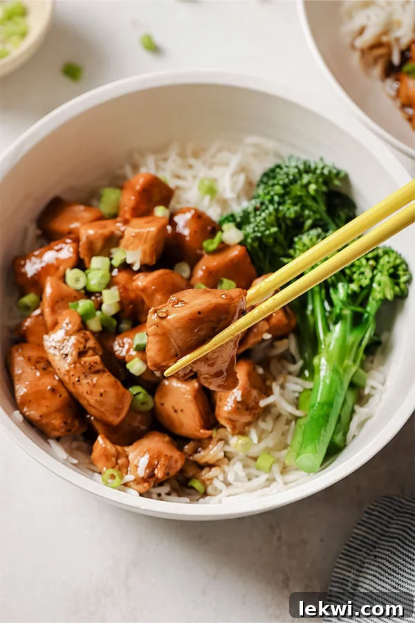 A vibrant and appealing bowl of healthy bourbon chicken served alongside fresh broccolini and delicate rice noodles, with chopsticks poised to take a delicious bite. The dish highlights the appetizing texture and rich color of the chicken.