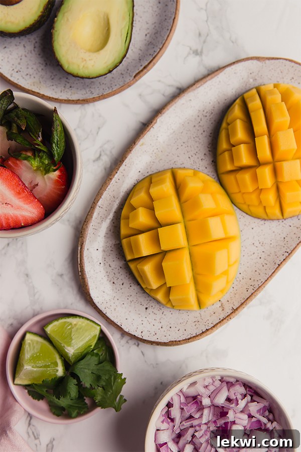A collection of fresh ingredients laid out on a wooden cutting board, including diced mango, sliced strawberries, a whole avocado, a red onion, a lime, and fresh cilantro, ready for making salsa.