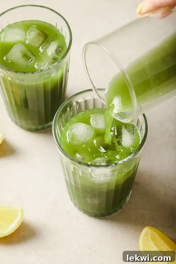 Matcha lemonade being poured from a pitcher into a glass filled with ice and lemon slices.