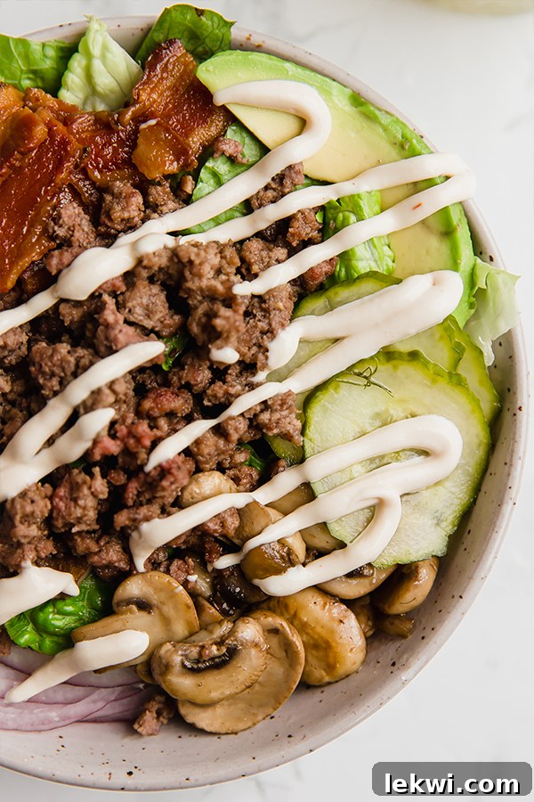 A burger bowl with lettuce, ground beef, pickles, avocado, and mushrooms with sauce drizzled on top. 