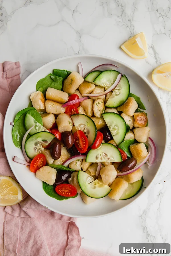 A white bowl filled with Greek gnocchi salad with a pink napkin next to it and extra lemon wedges, showcasing the fresh ingredients and vibrant colors.