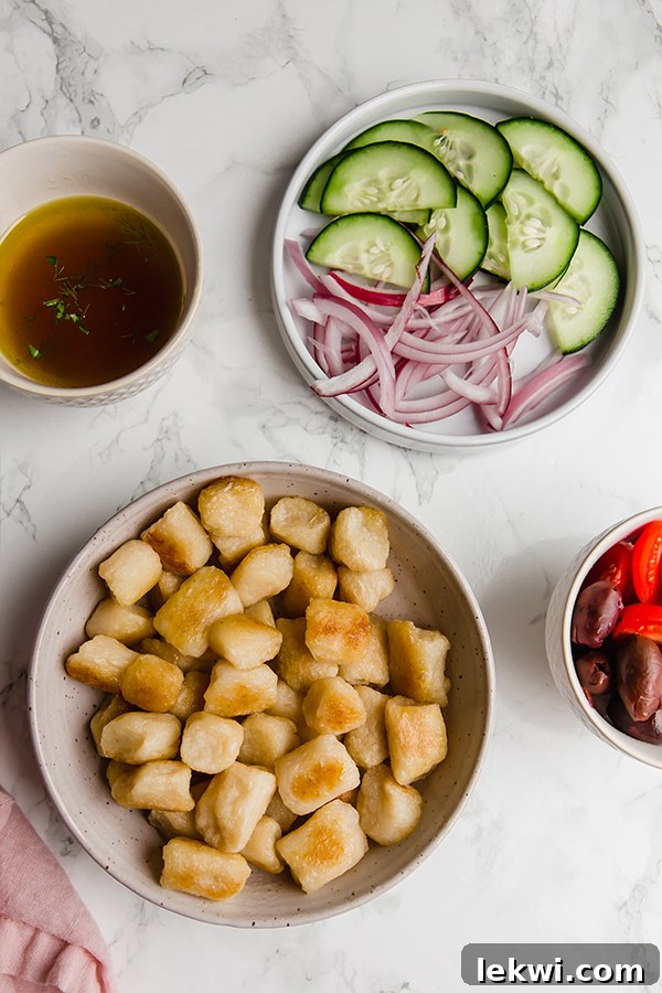 All the fresh ingredients needed to make Greek gnocchi salad, including cauliflower gnocchi, spinach, red onion, cucumber, tomatoes, and Kalamata olives, artfully arranged in bowls on a kitchen counter.