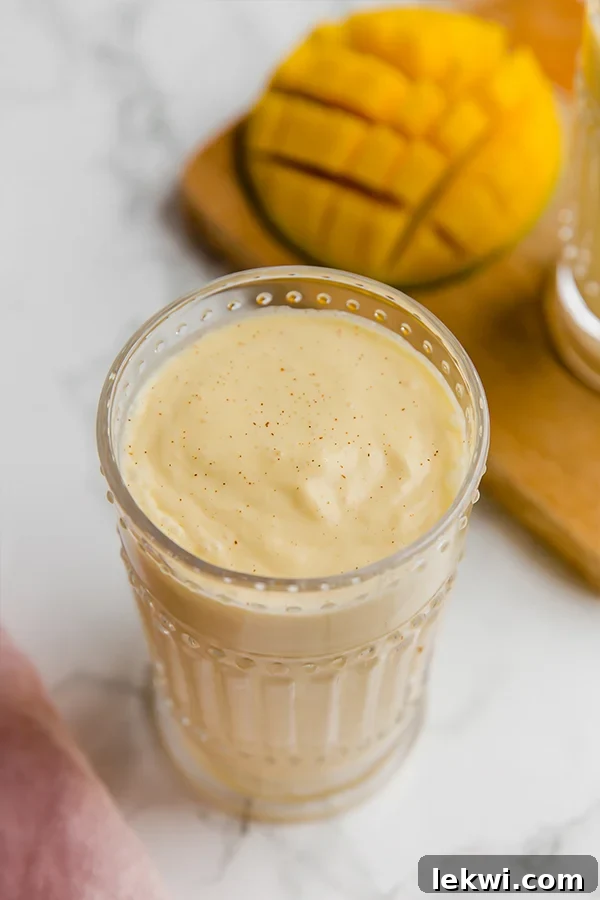 A glass filled with vibrant yellow mango lassi, garnished with a sprinkle of cinnamon and a fresh mango slice, sitting on a light-colored surface with natural light.