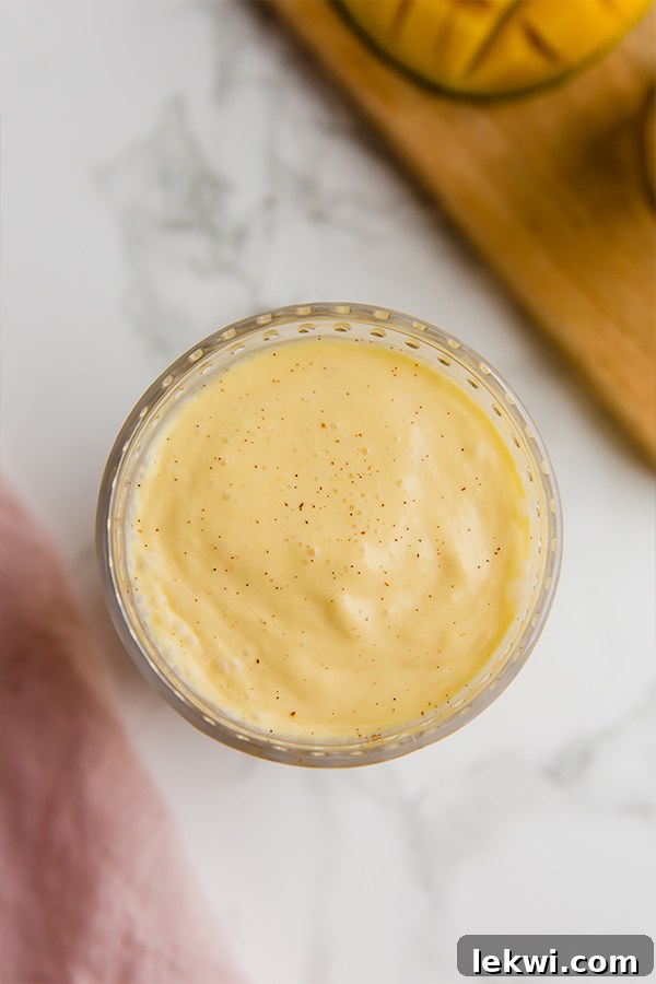 Two glasses of mango lassi on a table next to a bowl of fresh mango chunks and a cinnamon stick. The background is softly blurred.