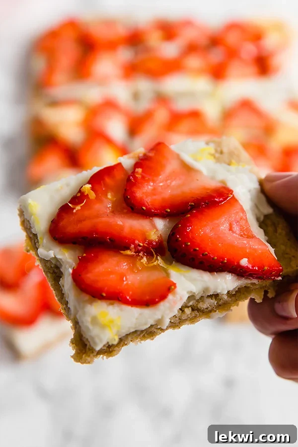 A stack of three strawberry lemon sugar cookie bars, beautifully layered with cookie, frosting, and fresh strawberries, sits invitingly on a simple white plate.