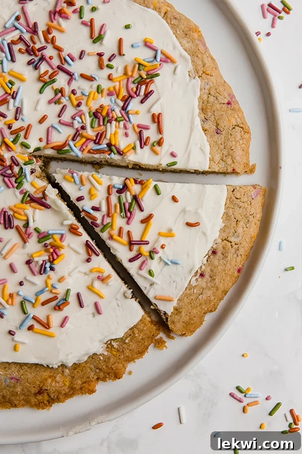 A white plate with a funfetti cookie pizza on it with a slice cut out but not yet removed.
