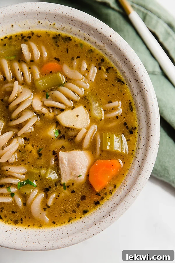 A steaming white bowl of classic chicken noodle soup, garnished with fresh herbs, invitingly placed on a wooden table.