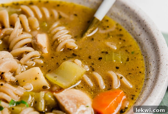 A white ceramic bowl filled with golden chicken noodle soup, with a spoon resting inside, ready to be enjoyed.