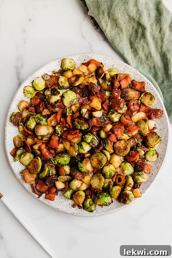 A side shot of the Bacon, Date & Apple Brussels Sprout Skillet on a white plate, accompanied by a clean linen napkin, highlighting the textures of the dish.