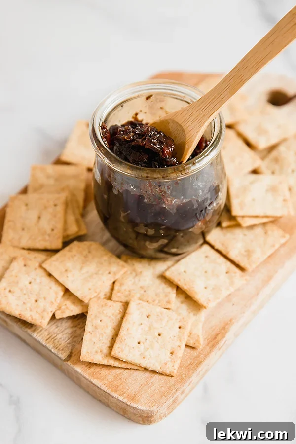 A beautifully arranged wooden board featuring an array of crackers and a glass jar filled with rich, homemade apple bacon jam, complete with a small wooden spoon resting in the jam.