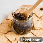 A wooden board filled with crackers and a glass jar with apple bacon jam and a wooden spoon in the jar of jam.