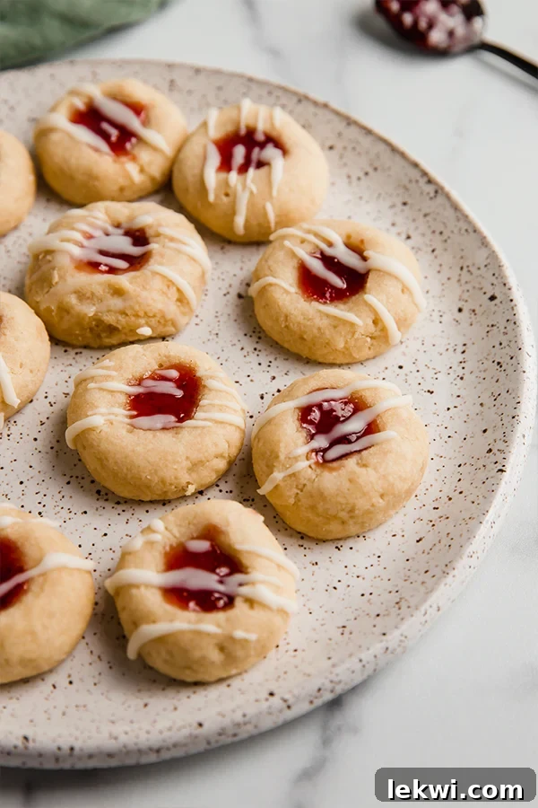 A beautifully arranged plate of gluten-free thumbprint cookies, each with a vibrant red jam filling and a delicate white drizzle.
