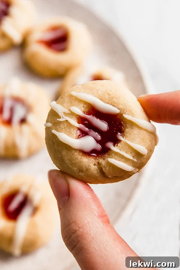 A close-up image of a hand holding a single gluten-free thumbprint cookie, showcasing its golden edge, red jam filling, and white coconut butter drizzle.