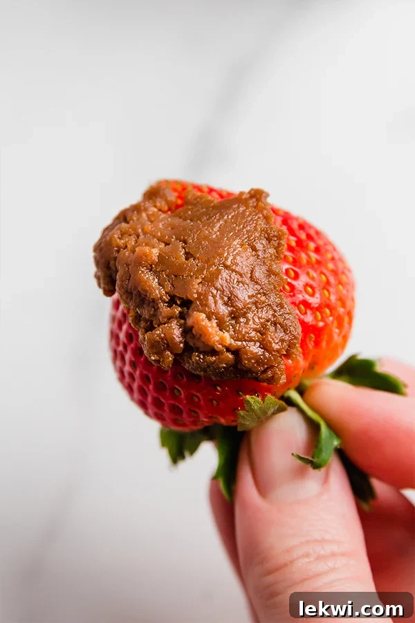 A vibrant red strawberry held with a dollop of creamy AIP cookie butter on its tip, illustrating a perfect pairing for a healthy snack.