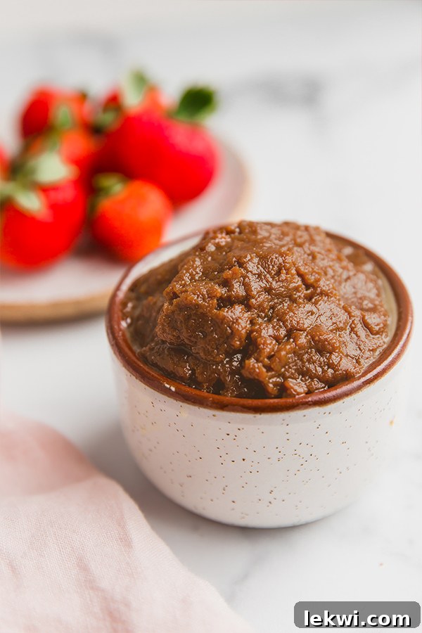 A charming close-up of AIP cookie butter in a small white cup, surrounded by fresh, vibrant red strawberries on a white plate, ready for a healthy snack.
