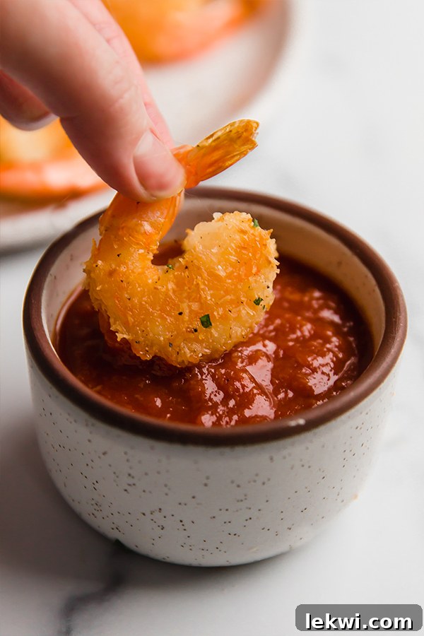 Oven-baked coconut shrimp being dipped into a vibrant red cocktail sauce.