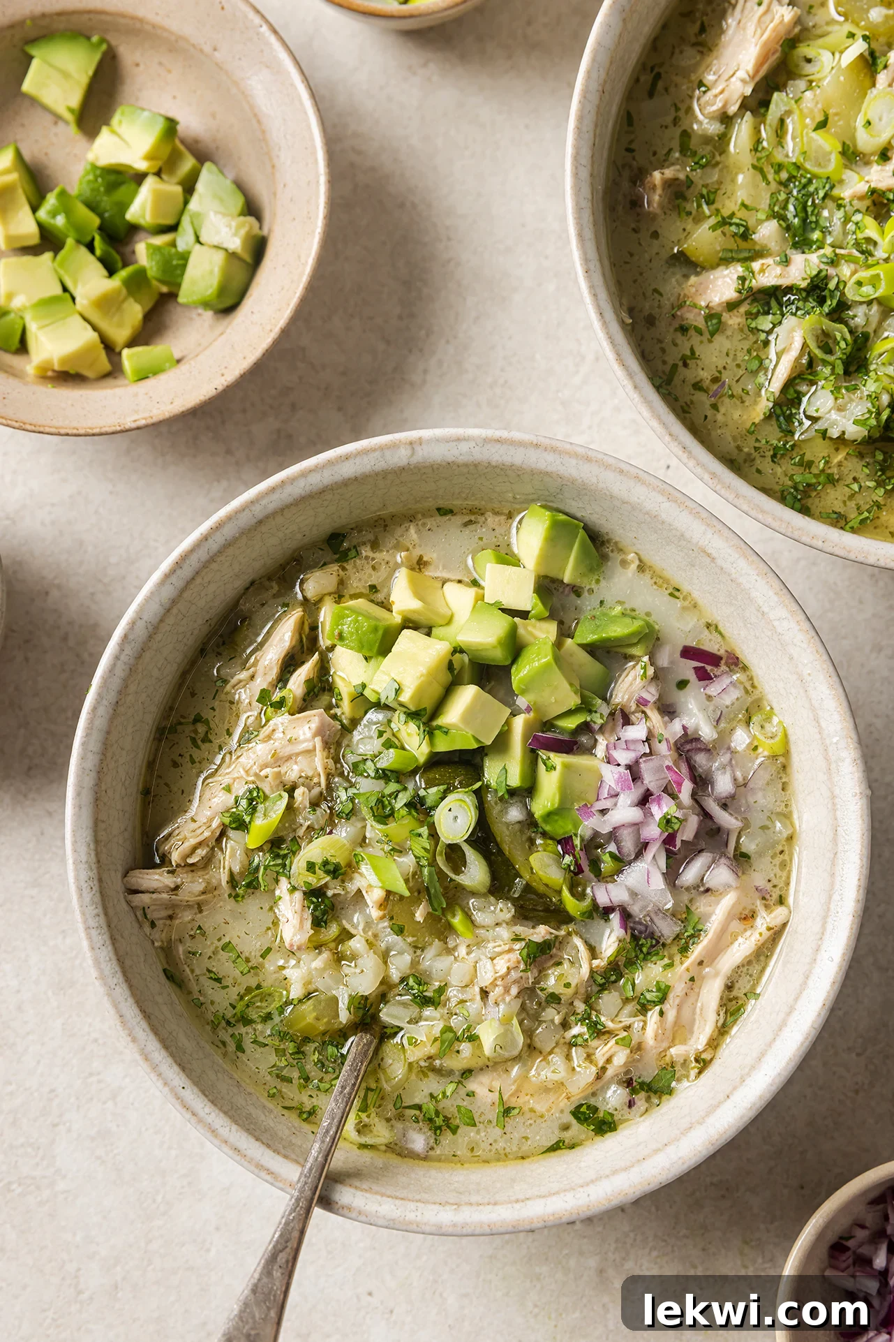 Three bowls of creamy White Chicken Chili, generously topped with fresh red onion and ripe avocado, presented on a rustic wooden surface.