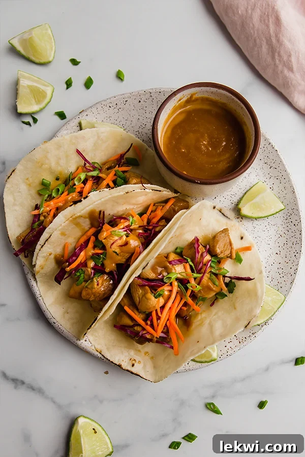 Three vibrant Thai chicken tacos arranged neatly on a white plate, showcasing the marinated chicken, colorful slaw, and a drizzle of almond butter sauce, with additional sauce served on the side in a small bowl.