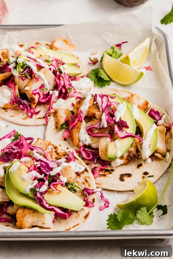 Three beautifully assembled fish tacos with vibrant slaw, avocado, and a creamy sauce, arranged on a serving tray with lime and cilantro.
