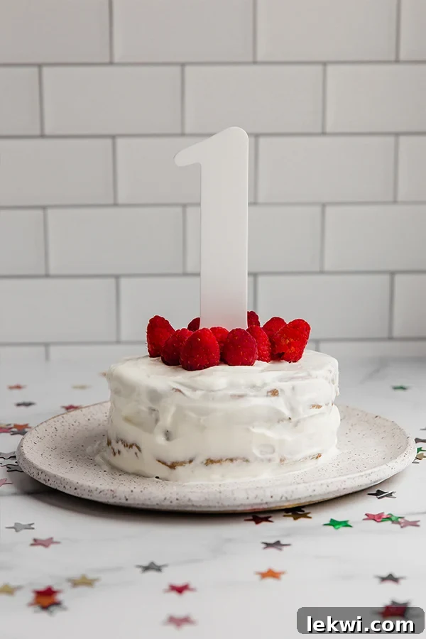 A baby joyfully interacting with a two-tier smash cake, decorated with raspberries and a 'one' cake topper, capturing a precious first birthday moment.