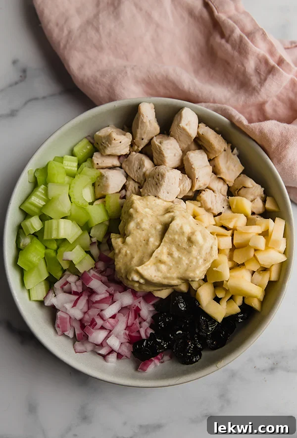 A large mixing bowl filled with cubed chicken, diced apple, chopped celery, red onion, and raisins, with a generous dollop of golden curry dressing on top, ready for tossing.