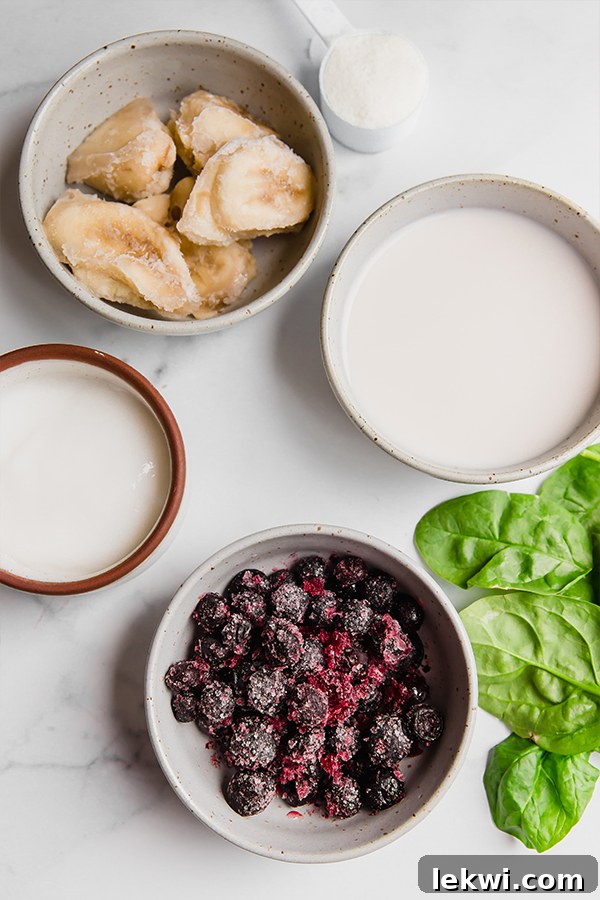 A vibrant flat lay showcases essential smoothie bowl ingredients: bright green spinach, slices of frozen banana, a bowl of frozen blueberries, and a spoon, all neatly arranged on a pristine white surface.