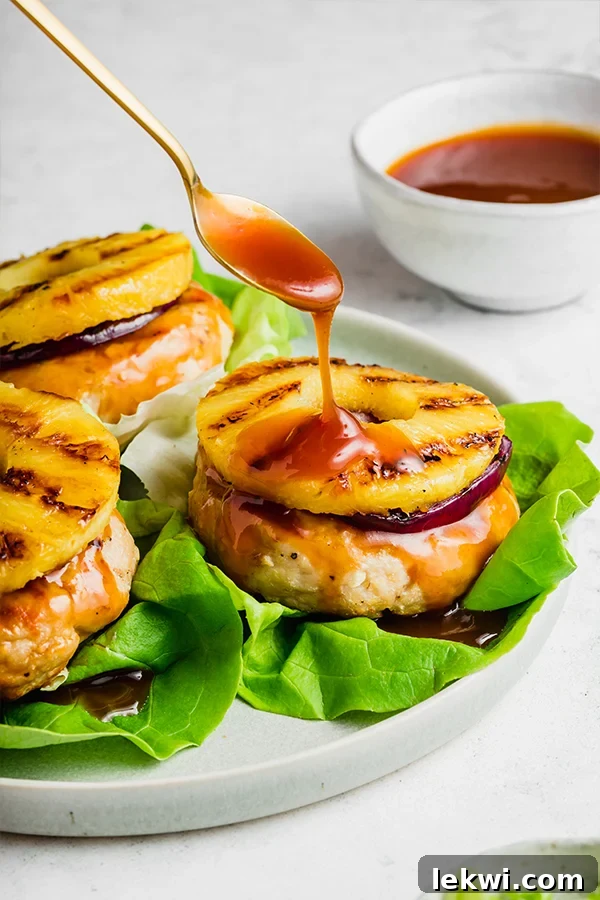 A close-up of a sweet and sour chicken burger being drizzled with sauce from a spoon, highlighting the rich, glossy texture of the homemade sauce and its appealing presentation.