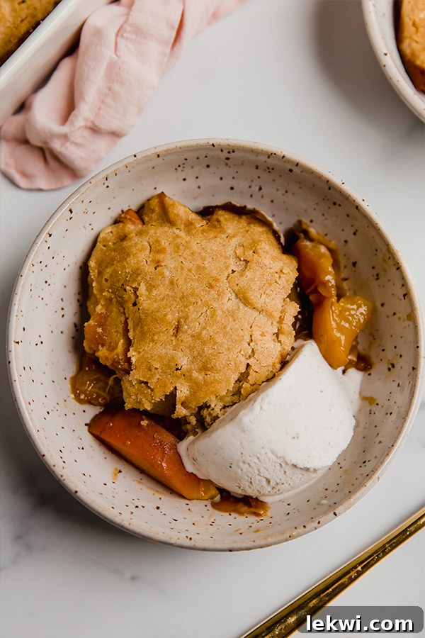 Top-down view of a freshly baked Paleo & AIP Peach Cobbler, showing a golden biscuit topping and a scoop of dairy-free vanilla ice cream melting on top.