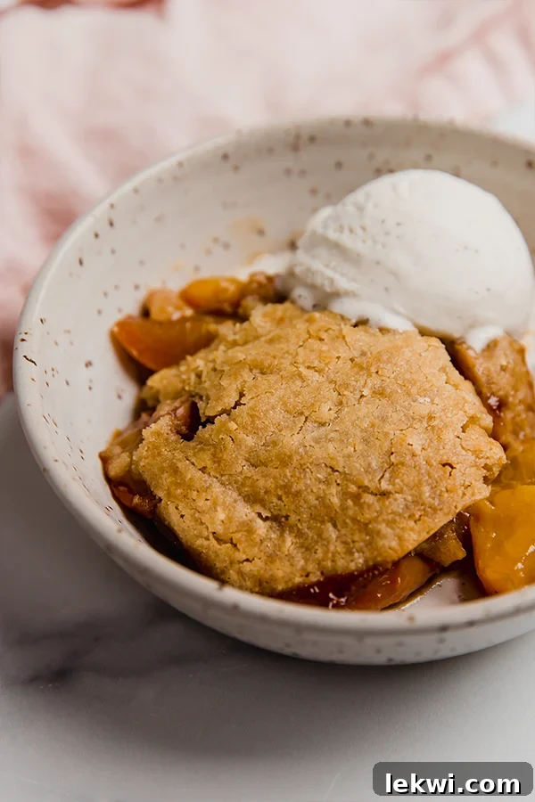 A serving of warm Paleo & AIP Peach Cobbler in a bowl, topped with a generous scoop of dairy-free ice cream that's beginning to melt, highlighting the golden biscuit topping.