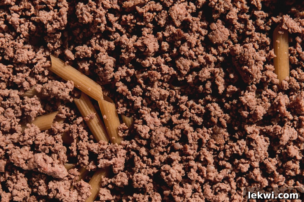 Cooked ground beef being added on top of the uncooked pasta in a baking dish.