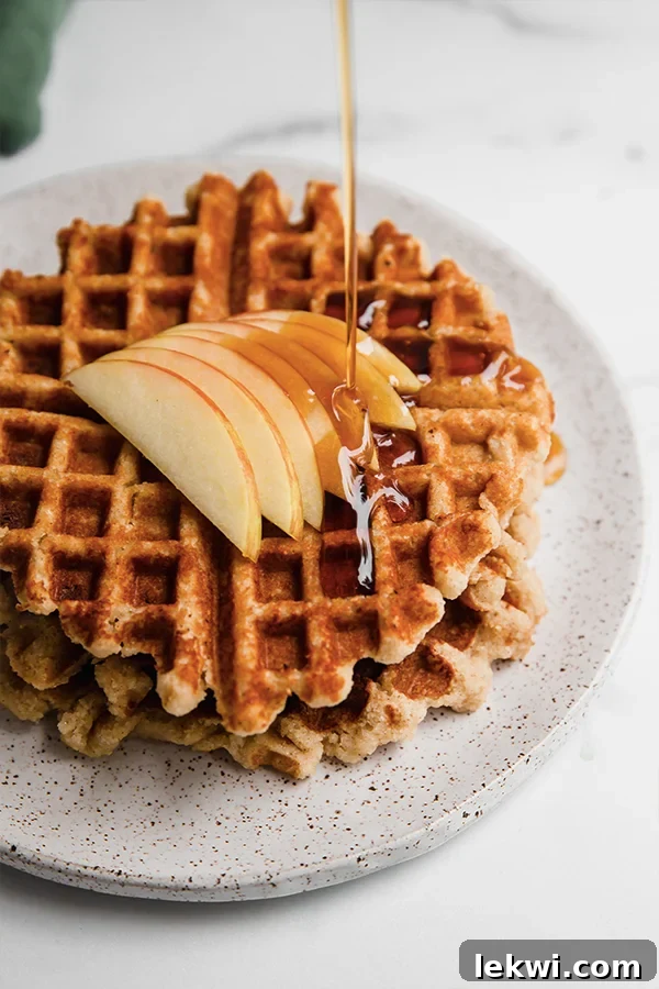 Syrup pouring over a stack of gluten-free apple waffles, garnished with apple slices.