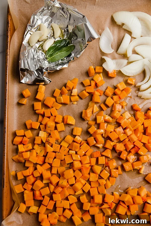 A sheet pan with raw sliced onion, cubed butternut squash, and a foil packet of garlic and sage, ready for roasting.